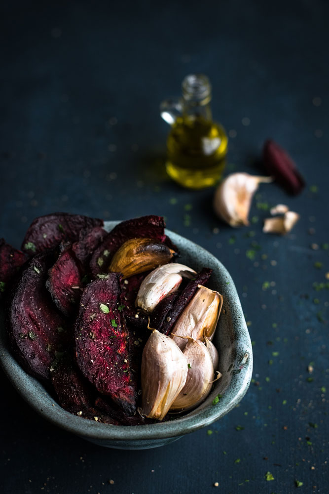 Bowl with roasted beets and garlic cloves in the foreground, with ingredients in the background